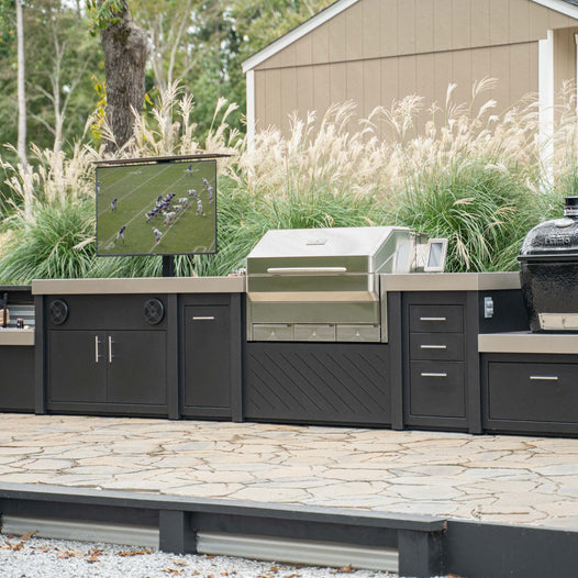 Stono Outdoor Living kitchen island, built-in gas grill, a Primo kamado grill, an outdoor refrigerator, an outdoor TV, a firewood rack, and a stainless steel sink with faucet.