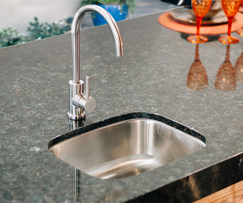 Stono Outdoor Living The TrueFlame 19" x 15" Stainless Steel Undermount Sink (TF-NK-19U) features a modern design with a curved faucet, set in polished dark granite. Orange glassware and a centerpiece in the background add charm—ideal for any outdoor kitchen.