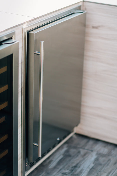 Stono Outdoor Living A close-up of the TrueFlame 24" 5.3 Cu. Ft. Deluxe Outdoor Rated Refrigerator (TF-RFR-24D-R) by TrueFlame, built into light wood cabinetry with a sleek stainless steel door and long vertical handle, next to a glass-fronted appliance on a wooden floor.
