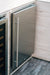 Stono Outdoor Living A close-up of the TrueFlame 24" 5.3 Cu. Ft. Deluxe Outdoor Rated Refrigerator (TF-RFR-24D-R) by TrueFlame, built into light wood cabinetry with a sleek stainless steel door and long vertical handle, next to a glass-fronted appliance on a wooden floor.