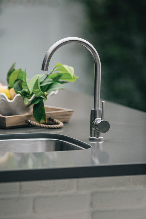 Stono Outdoor Living The TrueFlame TF-NK-19U 19" x 15" stainless steel undermount sink is set in a gray countertop, paired with a modern stainless steel faucet. In the background, a wooden tray displays lemons and green leaves.