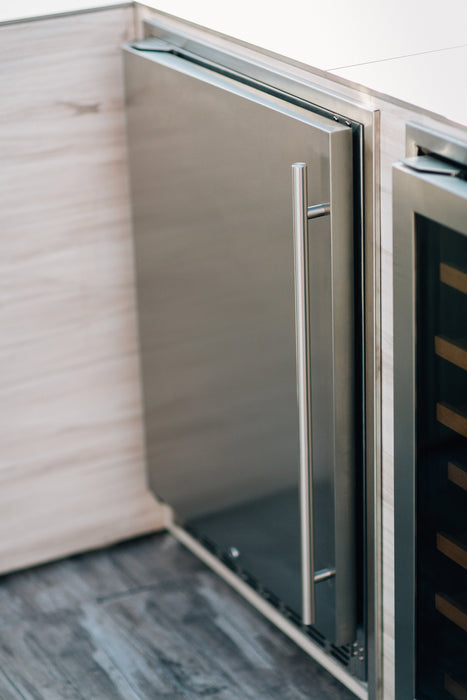 Stono Outdoor Living A close-up of the TrueFlame 24" 5.3 Cu. Ft. Deluxe Outdoor Rated Refrigerator (TF-RFR-24D-R) showcases its modern stainless steel design with a vertical handle, seamlessly integrated into light wood cabinetry beside a glass-door wine cooler.