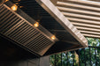 Stono Outdoor Living A close-up of the TrueFlame 36" Outdoor Vent Hood (TF-VH36-2) by TrueFlame, featuring built-in lights, installed in an outdoor kitchen under a slatted roof with sunlight and trees visible in the background.