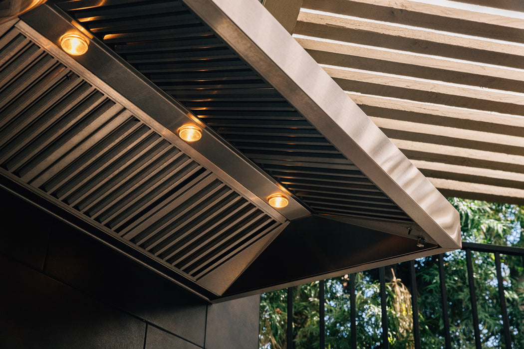 Stono Outdoor Living Close-up of the TrueFlame 36" Outdoor Vent Hood (TF-VH36-2) with built-in lights on, mounted under a slatted pergola. Sunlight and greenery are visible through the slats in the background.