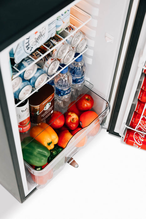 Stono Outdoor Living The TrueFlame 22" 4.1c Deluxe Outdoor Fridge (AMD-RFR-22D) is shown open in an outdoor kitchen, holding cans, bottled water, yogurt, apples, a green bell pepper, and an orange bell pepper in the door compartment.