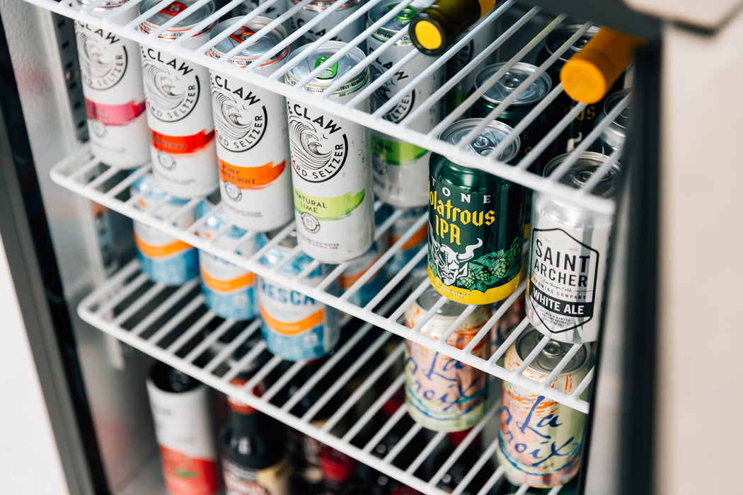Stono Outdoor Living The TrueFlame 24" 5.3 Cu. Ft. Deluxe Outdoor Rated Refrigerator (TF-RFR-24D-R) by TrueFlame, with a stainless steel door, showcases cans like White Claw, Saint Archer IPA, and LaCroix—combining stylish storage for any outdoor space.