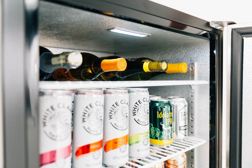 Stono Outdoor Living A TrueFlame 24" Outdoor Rated Refrigerator (Right Hinge) displays White Claw hard seltzer, an IPA beer can, and wine bottles neatly stored on its shelves.