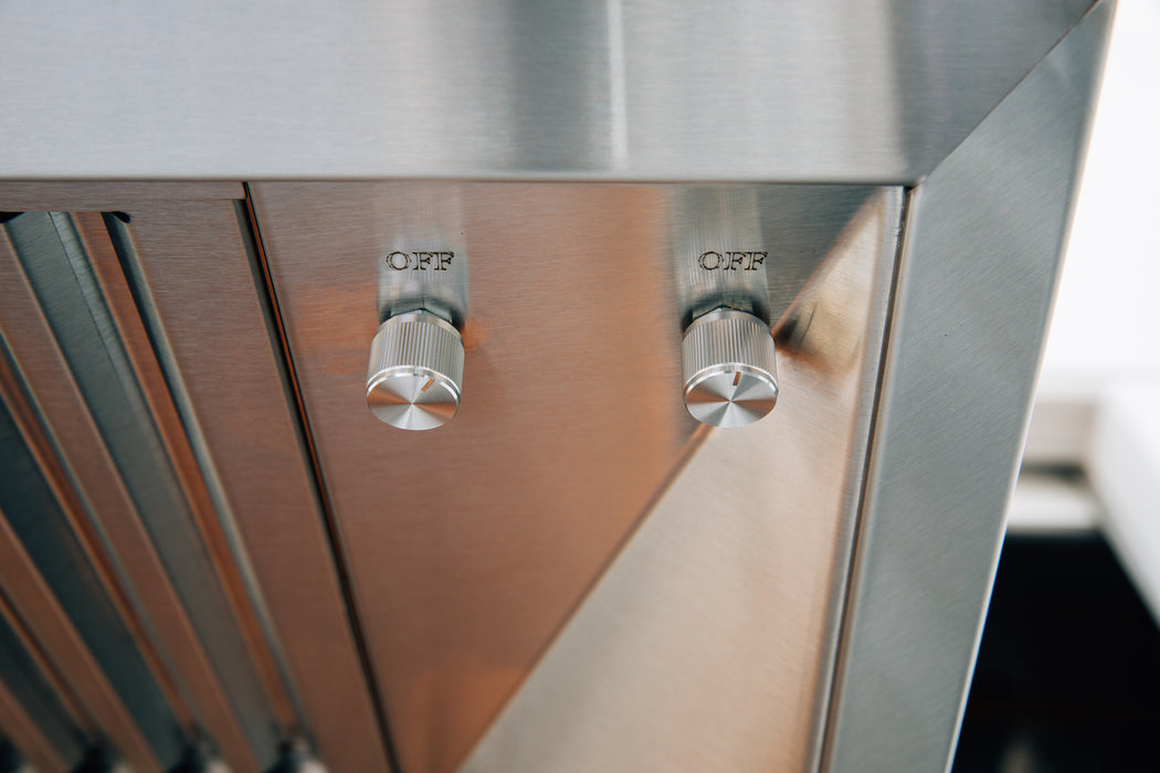 Stono Outdoor Living Close-up of two metallic control knobs labeled OFF on a TrueFlame 36" Outdoor Vent Hood (TF-VH36-2). The stainless steel knobs gleam as they reflect the surrounding light, highlighting the appliance's premium finish.
