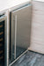 Stono Outdoor Living A TrueFlame 24" Outdoor Rated Refrigerator (5.1 Cu. Ft., Right Hinge, Model AMD-RFR-24S-B) is built into light wood cabinetry with a long vertical handle and sleek, modern design, sitting on a dark wood floor.