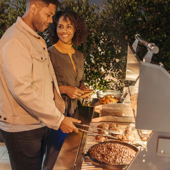 Stono Outdoor Living A man grills assorted meats on the American Made Grills Estate 30-Inch Natural Gas Grill (EST30-NG) while a woman smiles beside him in their green backyard, enjoying a pleasant evening together.