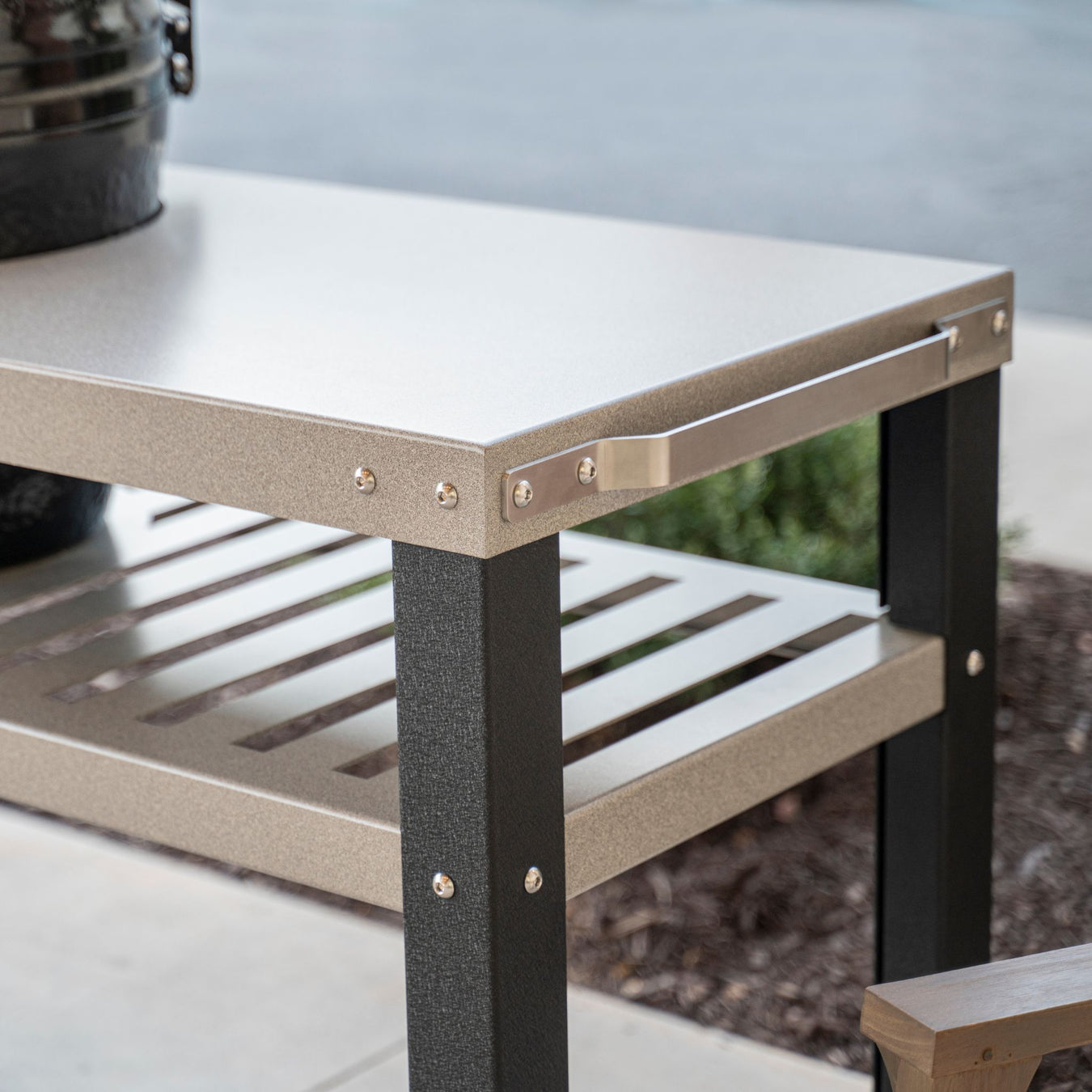 Stono Outdoor Living Outdoor metal table with a textured light gray top, black legs, a lower slatted shelf, and a silver handle on one side, placed on a patio next to mulch and a sidewalk.