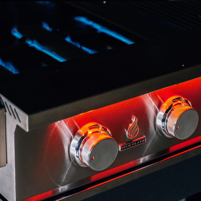 Stono Outdoor Living Close-up of a TrueFlame 32" 4-Burner Built-In Natural Gas Grill (TF32) with two orange-lit control knobs labeled TrueFlame and visible blue flames inside, showing the grill is on and heating.