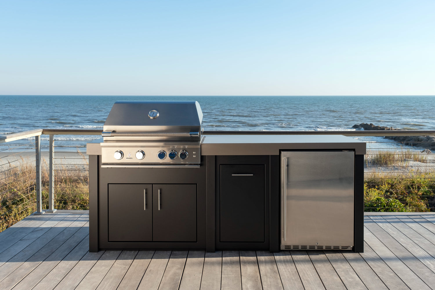 Stono Outdoor Living A modern outdoor kitchen with a grill, cabinets, and a refrigerator sits on a wooden deck overlooking a beach and the ocean under a clear sky.