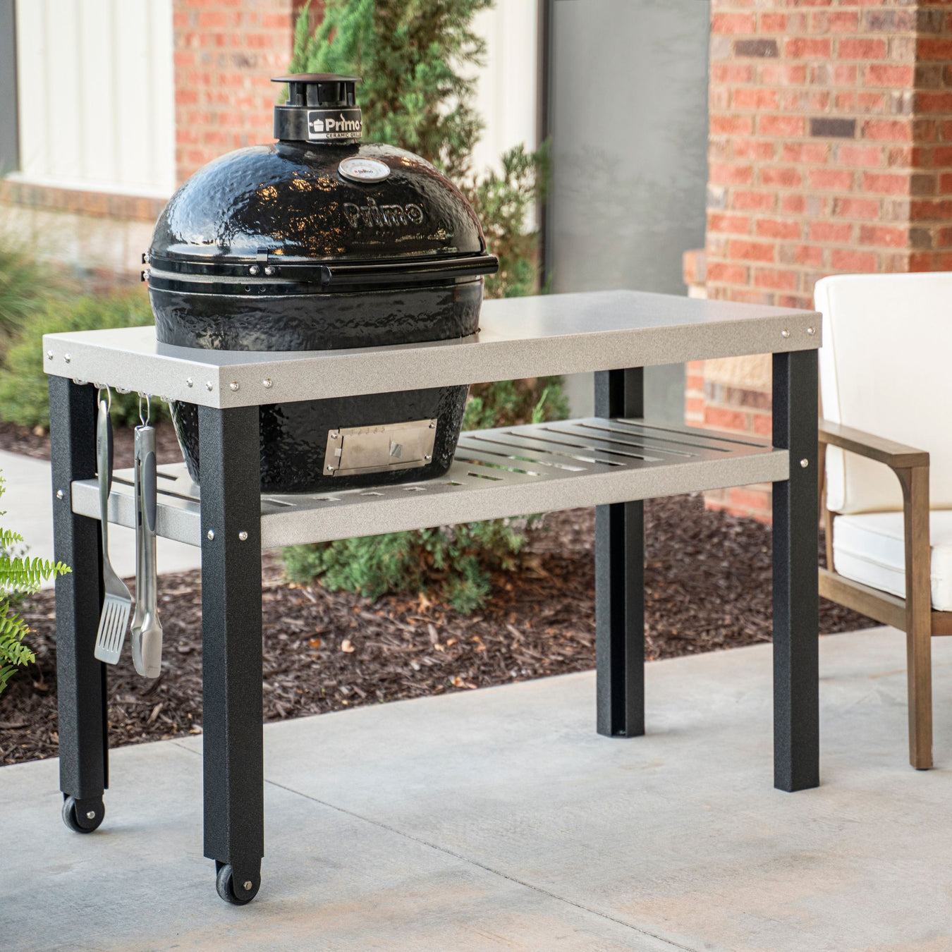 Stono Outdoor Living A black ceramic grill is set into a gray outdoor table with a lower metal shelf, cooking utensils hanging on the side, and a chair nearby, positioned on a patio with brick and plants in the background.