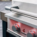Stono Outdoor Living Close-up of the TrueFlame 25" 3-Burner Built-In Propane Gas Grill (TF25L), featuring stainless steel construction, two red control knobs, the TrueFlame logo, and a textured handle on a tiled patio.