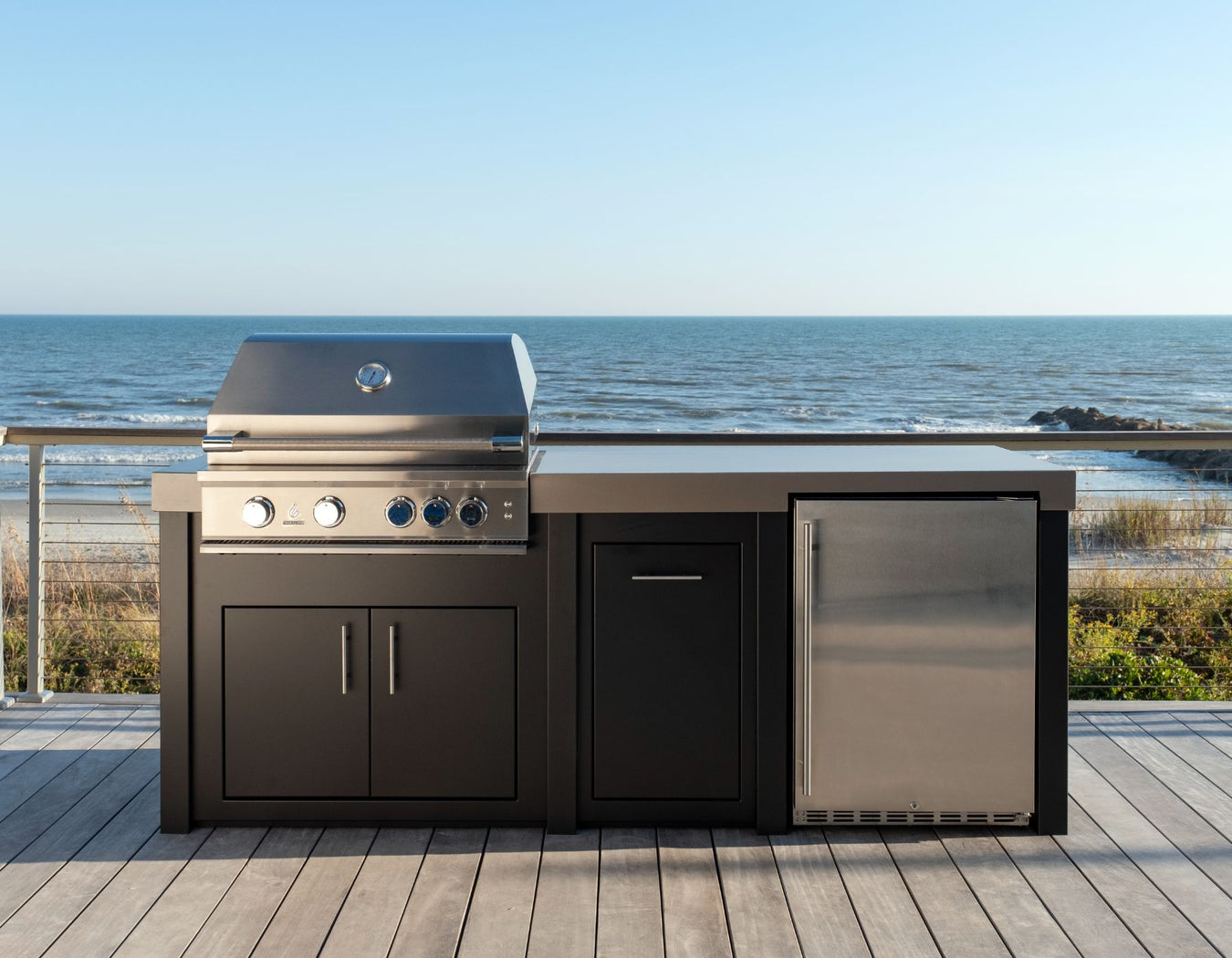 Stono Outdoor Living A modern outdoor kitchen with a grill, storage cabinets, and a refrigerator sits on a wooden deck overlooking the ocean under a clear blue sky.