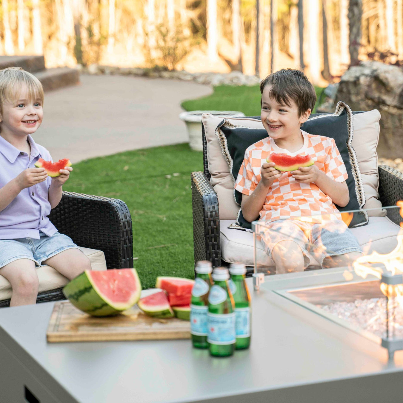 Stono Outdoor Living Two young children sit on outdoor chairs, smiling and eating watermelon. A sliced watermelon and bottles of sparkling water are on the table in front of them, with a fire pit and greenery in the background.