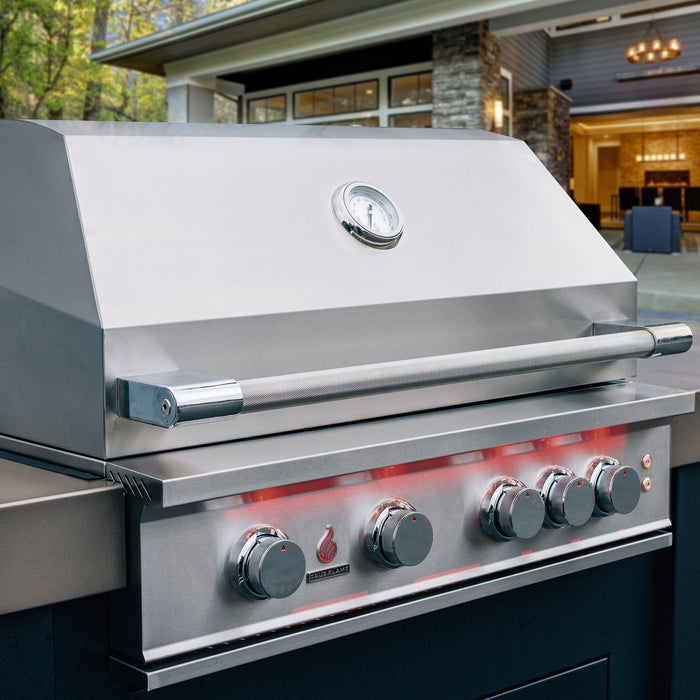 Stono Outdoor Living A close-up of the TrueFlame 32" 4-Burner Built-In Natural Gas Grill - TF32 shows its four control knobs, closed lid with built-in thermometer, and stainless steel finish, set on a patio with a house and trees in the background.