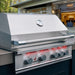 Stono Outdoor Living A close-up of the TrueFlame 32" 4-Burner Built-In Propane Gas Grill (TF32L) with a closed lid, temperature gauge, and four control knobs, set on a patio with a blurred house and trees in the background.