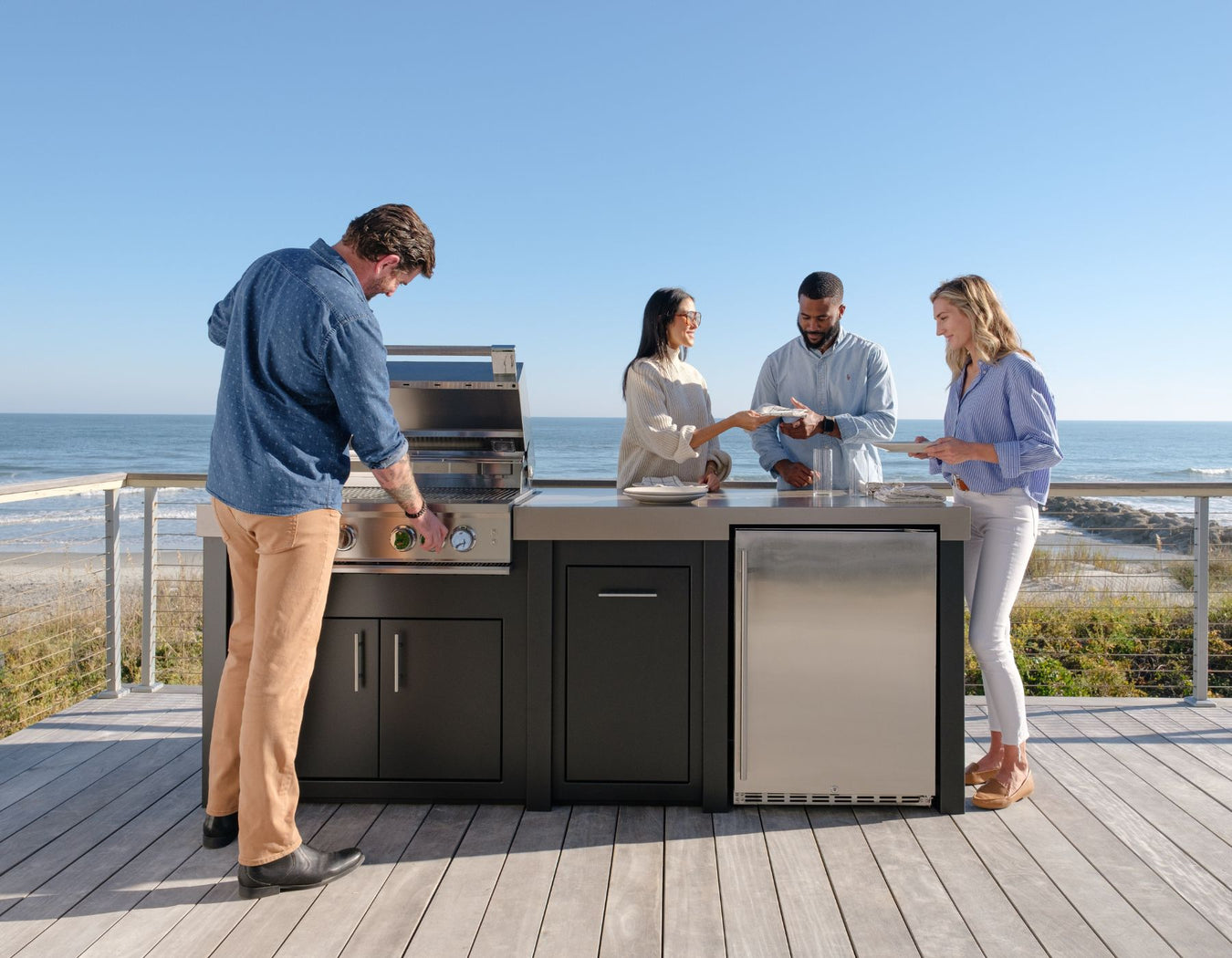 Stono Outdoor Living Four people are gathered around an outdoor kitchen on a wooden deck by the ocean. One person grills food while the others stand nearby, smiling and enjoying the sunny, seaside setting.
