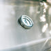 Stono Outdoor Living A close-up of a round temperature gauge on the sleek metallic surface of the American Made Grills Estate 30-Inch Grill (Natural Gas), with light and faint outdoor shadows reflecting in the background.