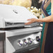 Stono Outdoor Living A woman in a sleeveless dress lifts the lid of a shiny Twin Eagles 30-Inch 2-Burner Built-In Natural Gas Grill with Sear Zone & Infrared Rotisserie Burner, with four knobs and white flowers in the background.