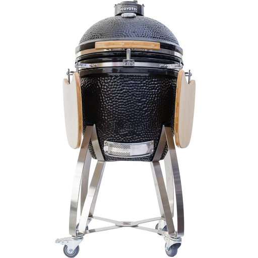 Stono Outdoor Living The Coyote Asado Smoker w/ Stand & Side Shelves (C1CHCS-FS) by Coyote is a black, egg-shaped kamado-style charcoal grill with a domed lid, wooden handles, foldable side shelves, and a wheeled metal stand for easy mobility.