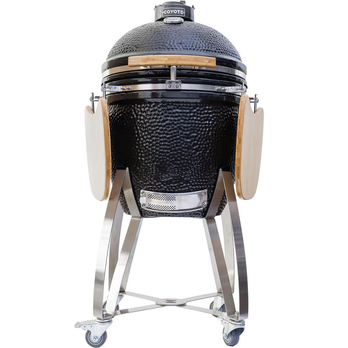 Stono Outdoor Living The Coyote Asado Smoker w/ Stand & Side Shelves (C1CHCS-FS) by Coyote is a black, egg-shaped kamado-style charcoal grill with a domed lid, wooden handles, foldable side shelves, and a wheeled metal stand for easy mobility.