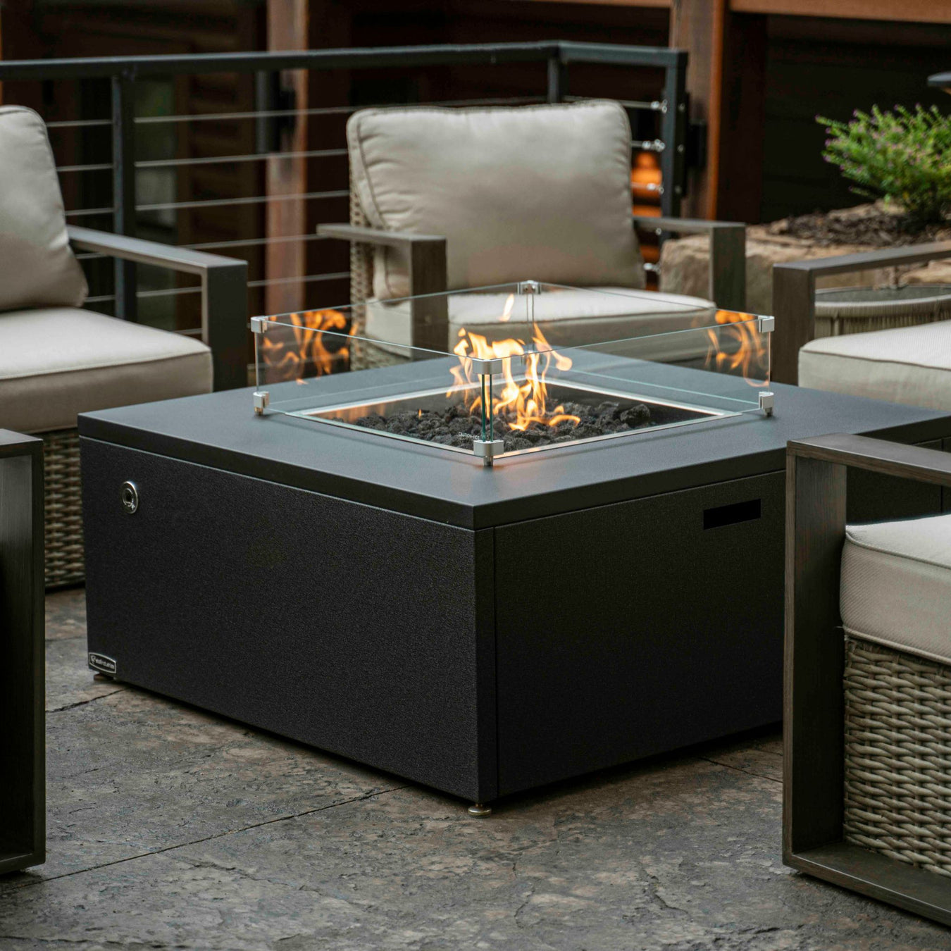 Stono Outdoor Living A modern square outdoor fire pit with a glass wind guard, surrounded by cushioned patio chairs on a stone patio. The fire pit is lit, creating a cozy ambiance.