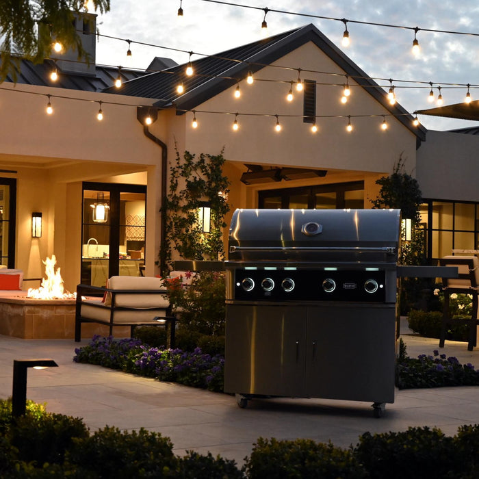 Stono Outdoor Living At dusk, an outdoor patio features the Wildfire Ranch Pro 36-in 3 Burner Black 304 SS Natural Gas Grill by Wildfire Outdoor Living, with string lights, lounge seating, a fire pit, potted plants, and a modern house with large windows in view.