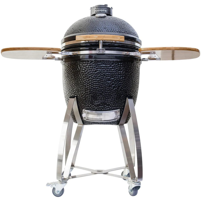 Stono Outdoor Living The Coyote Asado Smoker w/ Stand & Side Shelves (C1CHCS-FS) is a black ceramic kamado-style charcoal grill with a domed lid, wooden shelves, stainless steel legs, and wheels—ideal for outdoor cooking and shown on a white background.
