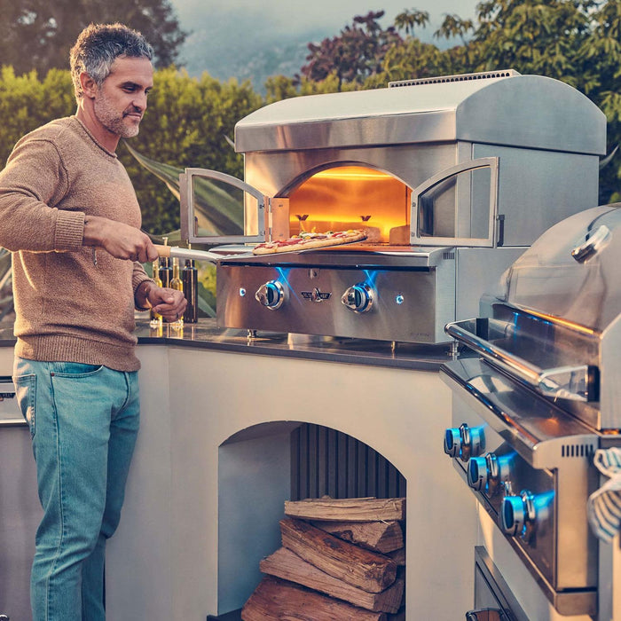 Stono Outdoor Living A man uses a pizza peel to remove a pizza from the Delta Heat 30-Inch Built-In Pizza Oven (Propane, DHPO30BI-L) by Delta Heat. A grill is beside the oven, firewood is stacked below, and trees fill the background.