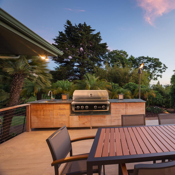 Stono Outdoor Living An outdoor kitchen featuring the Wildfire Ranch Pro 42" Gas Grill 304 Black SS Propane by Wildfire Outdoor Living, a sink, and wooden cabinets on a patio with a wooden dining set, surrounded by greenery and trees under a clear evening sky.