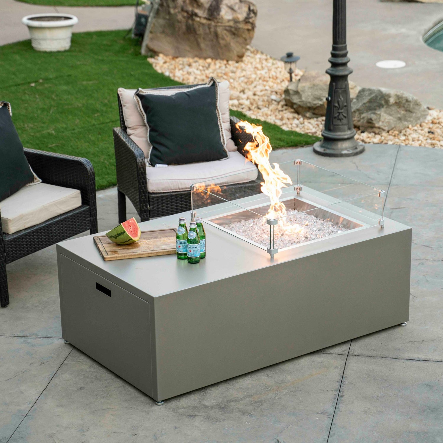 Stono Outdoor Living A modern outdoor patio scene with a rectangular fire pit table, three bottled drinks, and a plate of watermelon on top. Two cushioned chairs are nearby, and green grass and rocks are in the background.