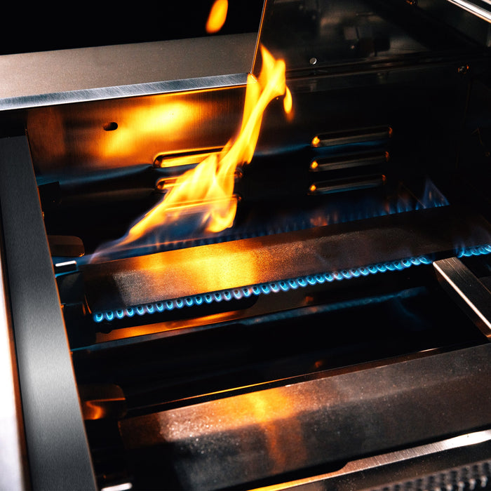 Stono Outdoor Living Close-up of the TrueFlame 25" 3-Burner Built-In Natural Gas Grill – TF25, featuring commercial-grade stainless steel, vivid blue flames, and a bright orange-yellow flare-up reflecting intense heat on the metal surfaces.