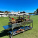 Stono Outdoor Living The Tagwood BBQ XL Double Argentine Santa Maria Wood Fire & Charcoal Grill (BBQ24SS) by Tagwood BBQ, loaded with meats and utensils, stands on grass with yachts docked behind—ready for open-fire grilling under a clear sky.