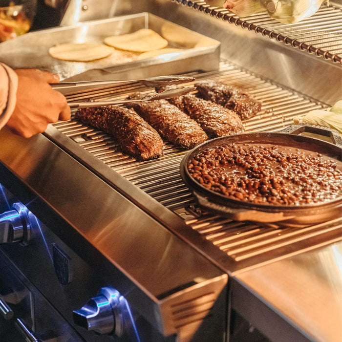 Stono Outdoor Living Someone uses tongs to grill steaks and heat a cast iron pan of baked beans on the American Made Grills Estate 30-Inch Grill (Natural Gas) by American Made Grills, with tortillas and vegetables cooking in the background.