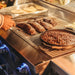 Stono Outdoor Living Someone uses tongs to grill steaks and heat a cast iron pan of baked beans on the American Made Grills Estate 30-Inch Grill (Natural Gas) by American Made Grills, with tortillas and vegetables cooking in the background.