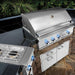 Stono Outdoor Living A Delta Heat 38-Inch 3-Burner Built-In Natural Gas Grill with infrared rotisserie burner (DHBQ38R-DN) sits in an outdoor kitchen featuring blue-lit knobs, a stone base, side burner, storage drawers, and a backdrop of greenery.