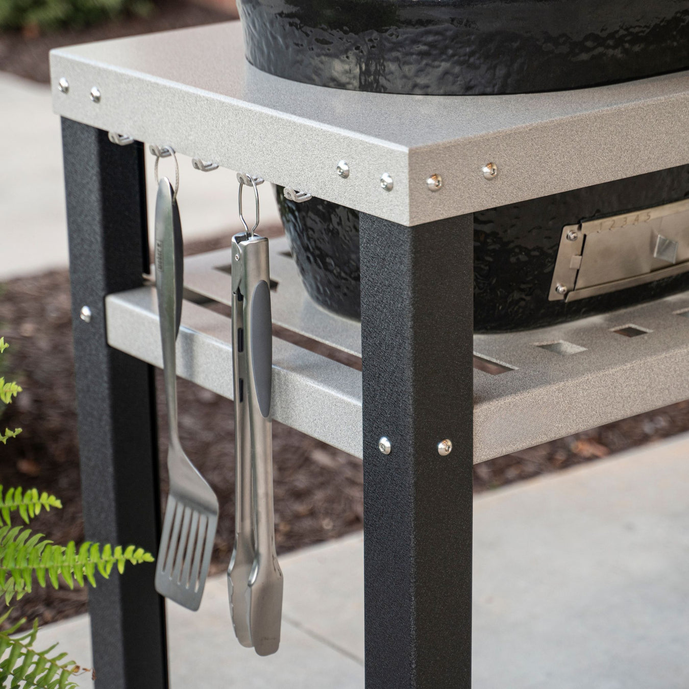Stono Outdoor Living Close-up of a metal grill table with a black grill on top. A spatula and tongs hang from hooks on the side. The scene is outdoors with blurred plants and pavement in the background.