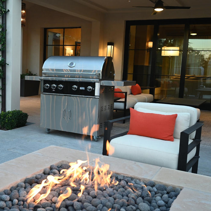 Stono Outdoor Living A modern patio with a Wildfire Outdoor Living Ranch Pro 36-in 3 Burner Black 304 SS Natural Gas Grill (WFPRO36G-BSS-NG), a fire pit with flames and black stones, and cushioned chairs with orange pillows by large windows.
