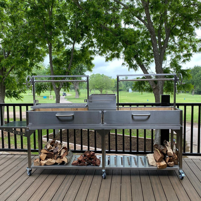 Stono Outdoor Living The Tagwood BBQ Double Argentine Santa Maria Wood Fire & Charcoal Grill (BBQ04SS) by Tagwood BBQ, featuring dual sections and overhead racks, stands on a deck with firewood below, set against a backdrop of trees and grassy parkland.