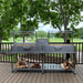 Stono Outdoor Living The Tagwood BBQ Double Argentine Santa Maria Wood Fire & Charcoal Grill (BBQ04SS) by Tagwood BBQ, featuring dual sections and overhead racks, stands on a deck with firewood below, set against a backdrop of trees and grassy parkland.
