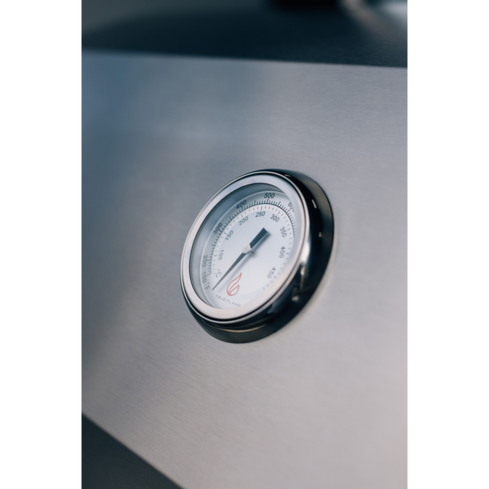 Stono Outdoor Living A close-up of the TrueFlame E Series 25" Stainless Steel 3-Burner Grill’s round metal thermometer, set in the stainless steel surface and displaying 100°F–700°F, with focus on the dial and a softly blurred background.
