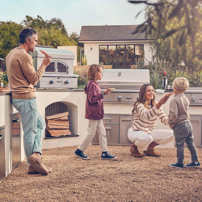 Stono Outdoor Living A family enjoys pizza outside; a man eats while a woman chats with two kids near an outdoor kitchen featuring the Delta Heat Freestanding Pizza Oven - Propane (DHPO30F-L) by Delta Heat, with a house and trees in the background.
