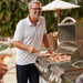 Stono Outdoor Living A smiling man in glasses and a white polo shirt grills shrimp and bread using the Blaze Premium LTE+ 32" 4-Burner Propane Grill with rear infrared burner and grill lights, surrounded by greenery and an umbrella.