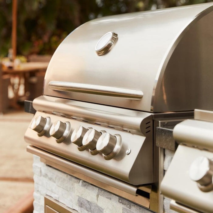Stono Outdoor Living A close-up of the Blaze Premium LTE+ 32" 4-Burner Propane Grill with rear infrared burner and grill lights, featuring premium searing rods and five knobs in a stone countertop, with a blurred patio and greenery behind.