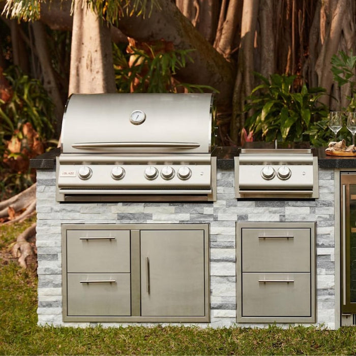 Stono Outdoor Living The Blaze Premium LTE+ 32" 4-Burner Propane Grill with rear infrared burner and grill lights is built into a stone counter with drawers and cabinets, surrounded by lush greenery and trees.