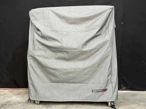 Stono Outdoor Living A large, gray Tagwood BBQ BBQ03SS/SI Cover - COVE03 with visible creases covers a rectangular object on wheels. The Tagwood BBQ logo patch is on the lower right corner. The background is dark.