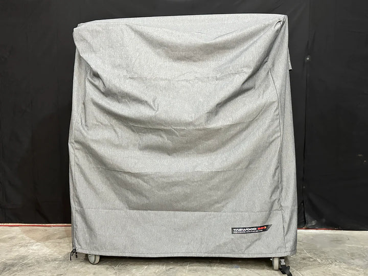Stono Outdoor Living A large, gray Tagwood BBQ BBQ03SS/SI Cover - COVE03 with visible creases covers a rectangular object on wheels. The Tagwood BBQ logo patch is on the lower right corner. The background is dark.